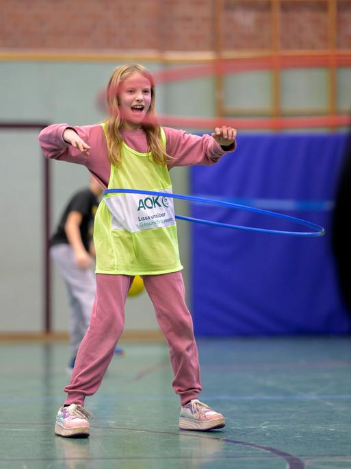 Ein blondes Mädchen im Grundschulalter steht in einer Turnhalle, trägt ein gelbes Leibchen über dem rosafarbenen Sportanzug und schwingt einen Hula-Hoop-Reifen um ihren Bauch. Ein blondes Mädchen im Grundschulalter steht in einer Turnhalle, trägt ein gelbes Leibchen über dem rosafarbenen Sportanzug und schwingt einen Hula-Hoop-Reifen um ihren Bauch.