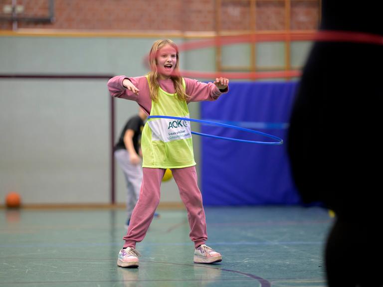 Ein blondes Mädchen im Grundschulalter steht in einer Turnhalle, trägt ein gelbes Leibchen über dem rosafarbenen Sportanzug und schwingt einen Hula-Hoop-Reifen um ihren Bauch. Ein blondes Mädchen im Grundschulalter steht in einer Turnhalle, trägt ein gelbes Leibchen über dem rosafarbenen Sportanzug und schwingt einen Hula-Hoop-Reifen um ihren Bauch.