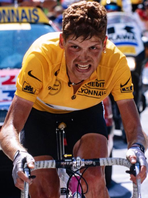Jan Ullrich fährt bei der Tour de France 1997 im Gelben Trikot des Gesamtführenden bergan. Die Kamera nimmt ihn von vorn auf, sein Geischt ist gezeichnet von der Anstrengung. Im Hintergrund unscharf ein Begleitwagen und -motorräder sowie Zuschauer.
