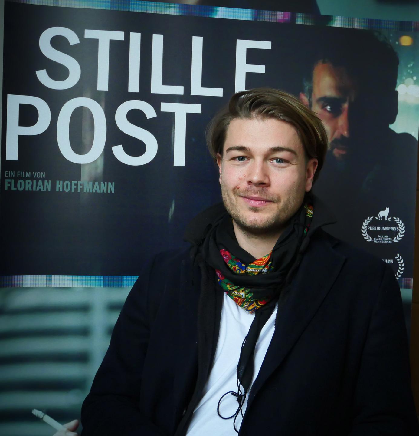 Der Regisseur Florian Hoffmann, aufgenommen in Berlin-Kreuzberg. Er steht vor dem Plakat seines Films "Stille Post". Die Haare trägt er zurückgekämmt und einen Drei-Tage-Bart.