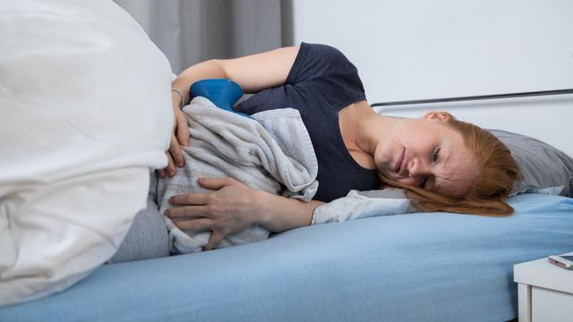 Eine junge Frau liegt in einem Bett. Sie hat eine Wärm-Flasche auf dem Bauch. 
