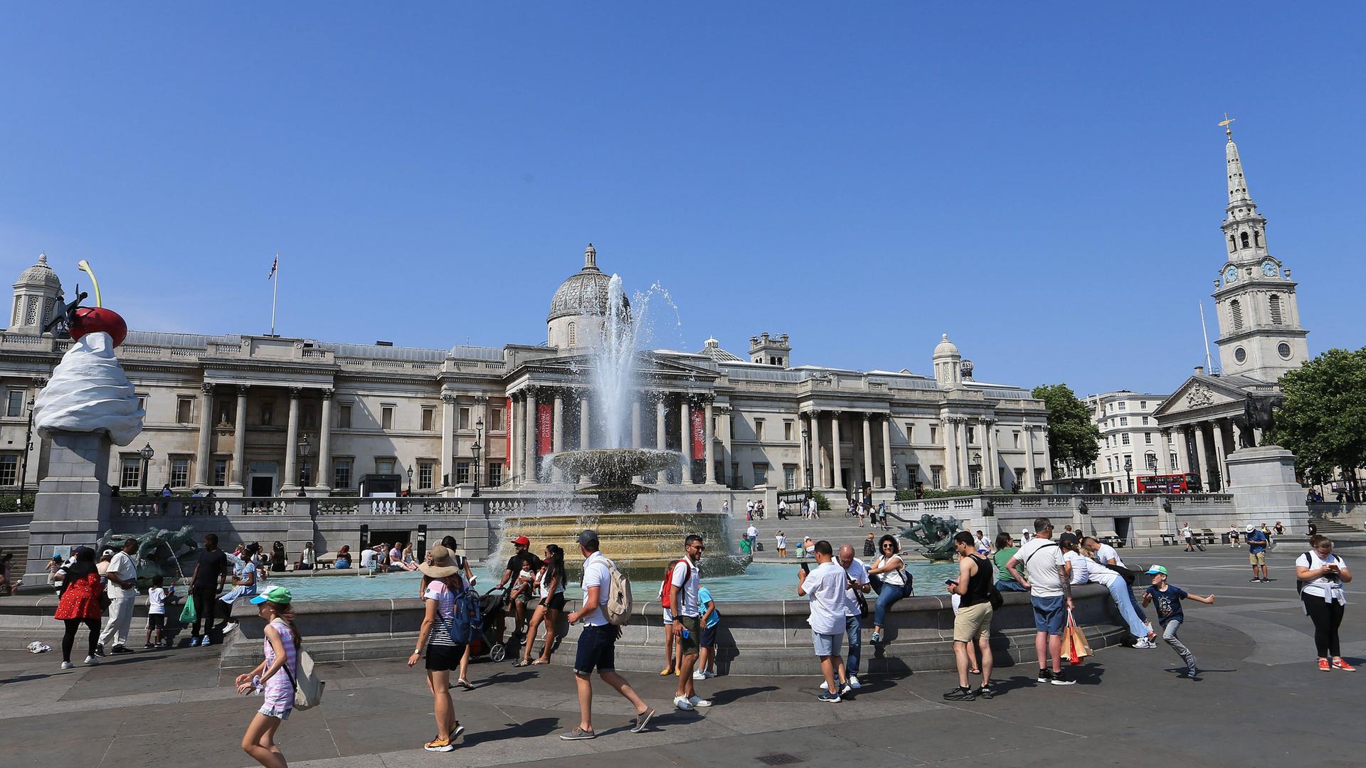 Menschen laufen bei strahlendem Sonnenschein und Hitze am Brunnen auf dem Trafalgar Aquare in London vorbei. Menschen laufen bei strahlendem Sonnenschein und Hitze am Brunnen auf dem Trafalgar Aquare in London vorbei.