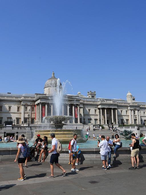 Menschen laufen bei strahlendem Sonnenschein und Hitze am Brunnen auf dem Trafalgar Aquare in London vorbei.