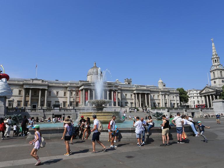 Menschen laufen bei strahlendem Sonnenschein und Hitze am Brunnen auf dem Trafalgar Aquare in London vorbei.