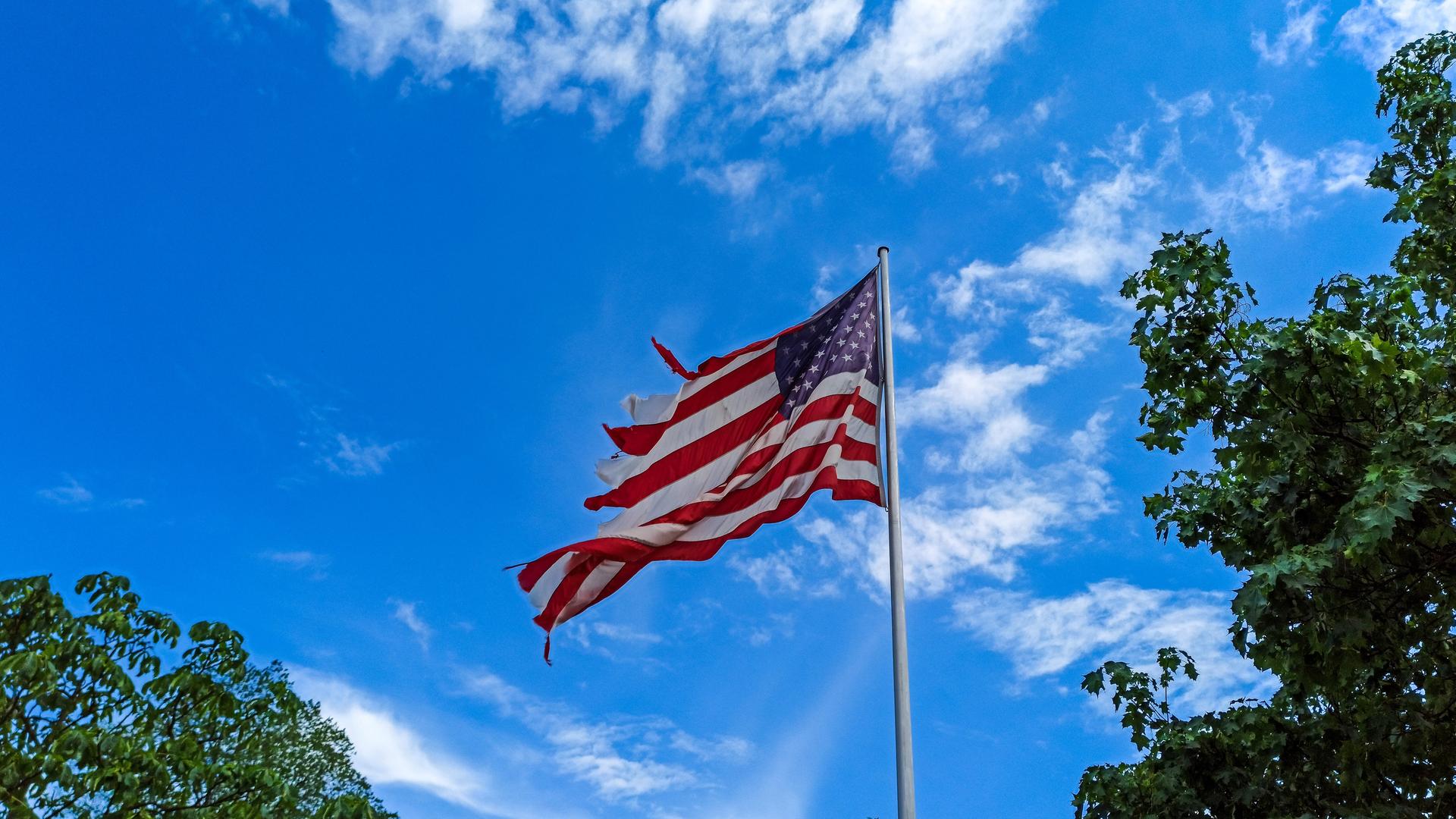 Eine im Wind wehende Flagge der Vereinigten Staaten von Amerika (USA) in einer zerfledderten Ausführung als Symbolbild für einen desolaten Zustand vor blauem, licht bewölkten Himmel. 