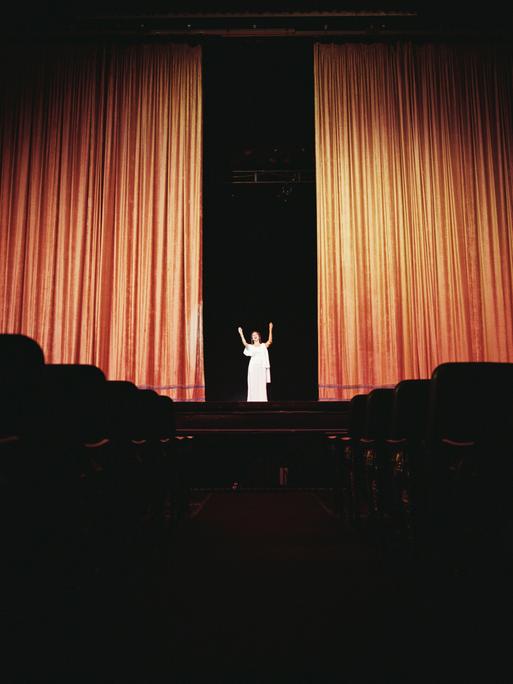 Auf einer Bühne geht ein Vorhang auf. In der Lücke dazwischen steht eine Frau in weißer Kleidung.