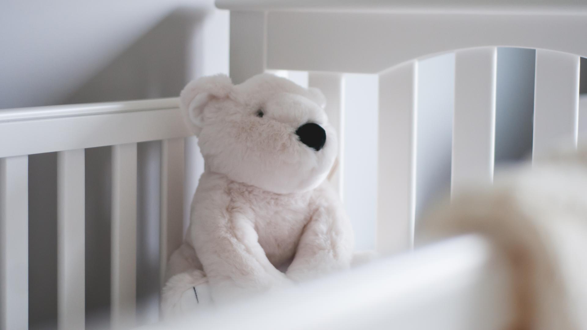 Ein weißer Teddybär sitzt ein einem weißen Kinderbett.