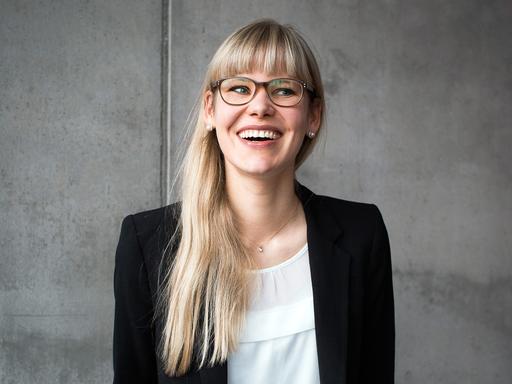 Die Psychologie-Professorin Eva Asselmann steht vor einer grauen Betonwand und guckt lachend zur Seite aus dem Bild hinaus.