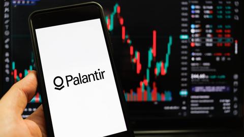 Das Palantir-Logo auf einem Smartphone-Bildschirm vor einem weiteren Bildschirm, der Datenanalyse-Kurven zeigt