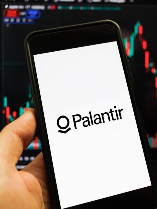 Das Palantir-Logo auf einem Smartphone-Bildschirm vor einem weiteren Bildschirm, der Datenanalyse-Kurven zeigt