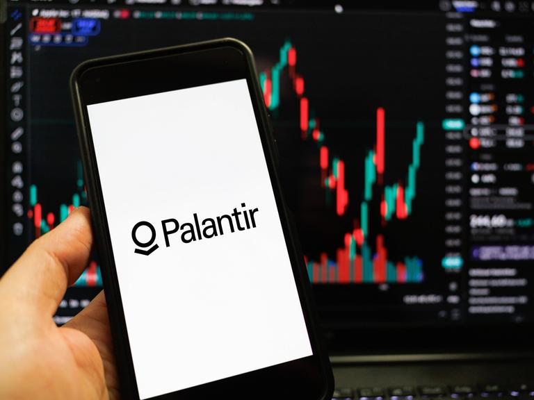 Das Palantir-Logo auf einem Smartphone-Bildschirm vor einem weiteren Bildschirm, der Datenanalyse-Kurven zeigt