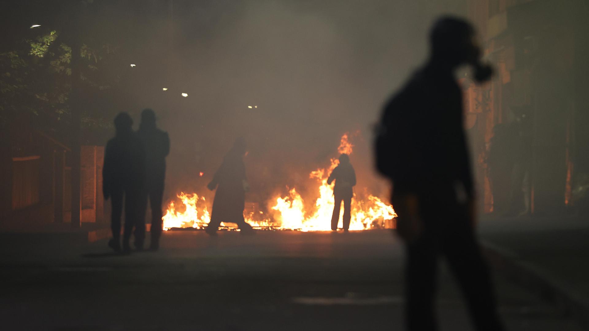 Ein Feuer in einer Stadt. Vor stehen Menschen, eine Person trägt eine Gasmaske. Es sind Proteste gegen die Regierung in Georgiens Hauptstadt Tiflis.