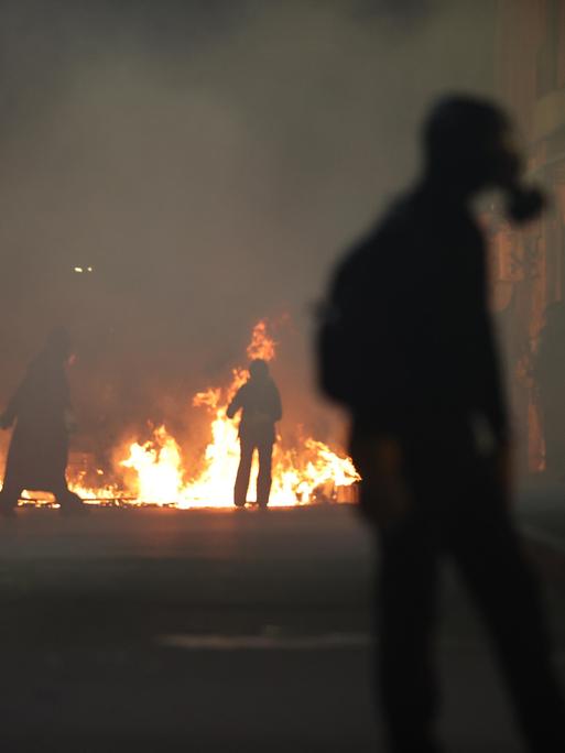 Ein Feuer in einer Stadt. Vor stehen Menschen, eine Person trägt eine Gasmaske. Es sind Proteste gegen die Regierung in Georgiens Hauptstadt Tiflis.