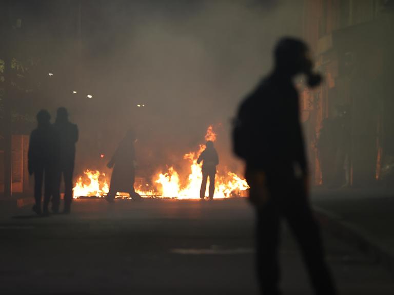Ein Feuer in einer Stadt. Vor stehen Menschen, eine Person trägt eine Gasmaske. Es sind Proteste gegen die Regierung in Georgiens Hauptstadt Tiflis.