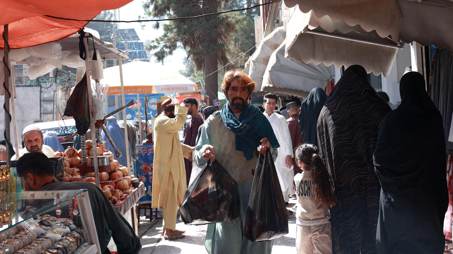 Ein Mann trägt Einkäufe auf einem Markt in Herat, daneben sind verhüllte Frauen und ein Mädchen zu sehen. Ein Mann trägt Einkäufe auf einem Markt in Herat, daneben sind verhüllte Frauen und ein Mädchen zu sehen.