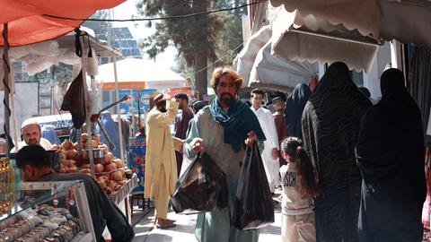 Ein Mann trägt Einkäufe auf einem Markt in Herat, daneben sind verhüllte Frauen und ein Mädchen zu sehen.