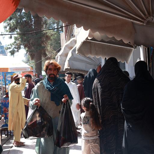 Ein Mann trägt Einkäufe auf einem Markt in Herat, daneben sind verhüllte Frauen und ein Mädchen zu sehen.