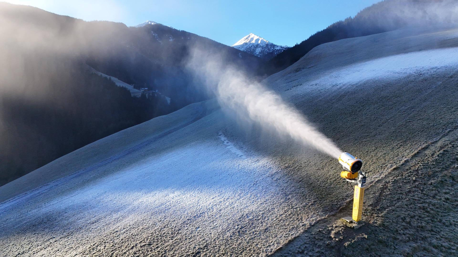 Das Bild zeigt einen Bergabhang ohne echten Schnee. Eine gelbe Schneekanone schießt weißen Kunstschnee auf die abschüssige Wiese.  