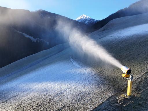 Das Bild zeigt einen Bergabhang ohne echten Schnee. Eine gelbe Schneekanone schießt weißen Kunstschnee auf die abschüssige Wiese.  