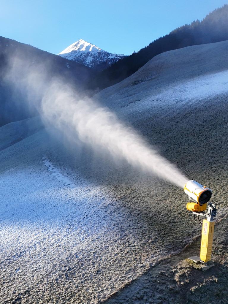 Das Bild zeigt einen Bergabhang ohne echten Schnee. Eine gelbe Schneekanone schießt weißen Kunstschnee auf die abschüssige Wiese.  