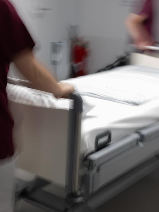 Eine Krankenpflegerin und ein Krankenpfleger schieben ein leeres Krankenbett durch den Gang einer Station eines Krankenhauses.