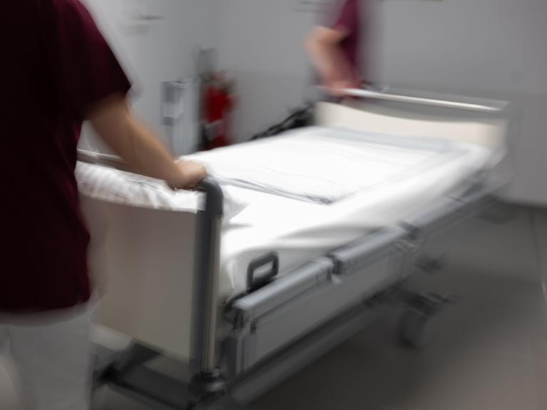 Eine Krankenpflegerin und ein Krankenpfleger schieben ein leeres Krankenbett durch den Gang einer Station eines Krankenhauses.