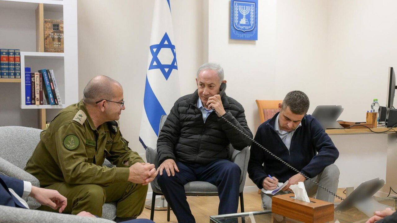 Benjamin Netanjahu hält einen Telefonhörer, während er mit anderen Männern vor einer israelischen Fahne zusammensitzt.