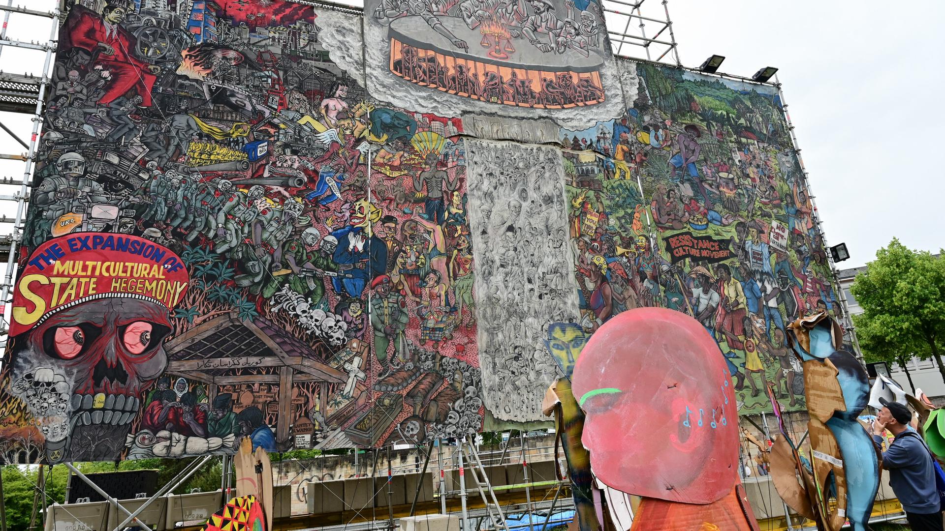 Auf dem etwa 10 x 10 Meter großen Bild der indonesischen Künstlergruppe Taring Padi ist ein Gewimmel verschiedenster Figuren zu sehen, links unten ein Totenkopf und auf dessen Kopfbedeckung die Aufschrift "The Expansion of Multicultural State Hegemony".