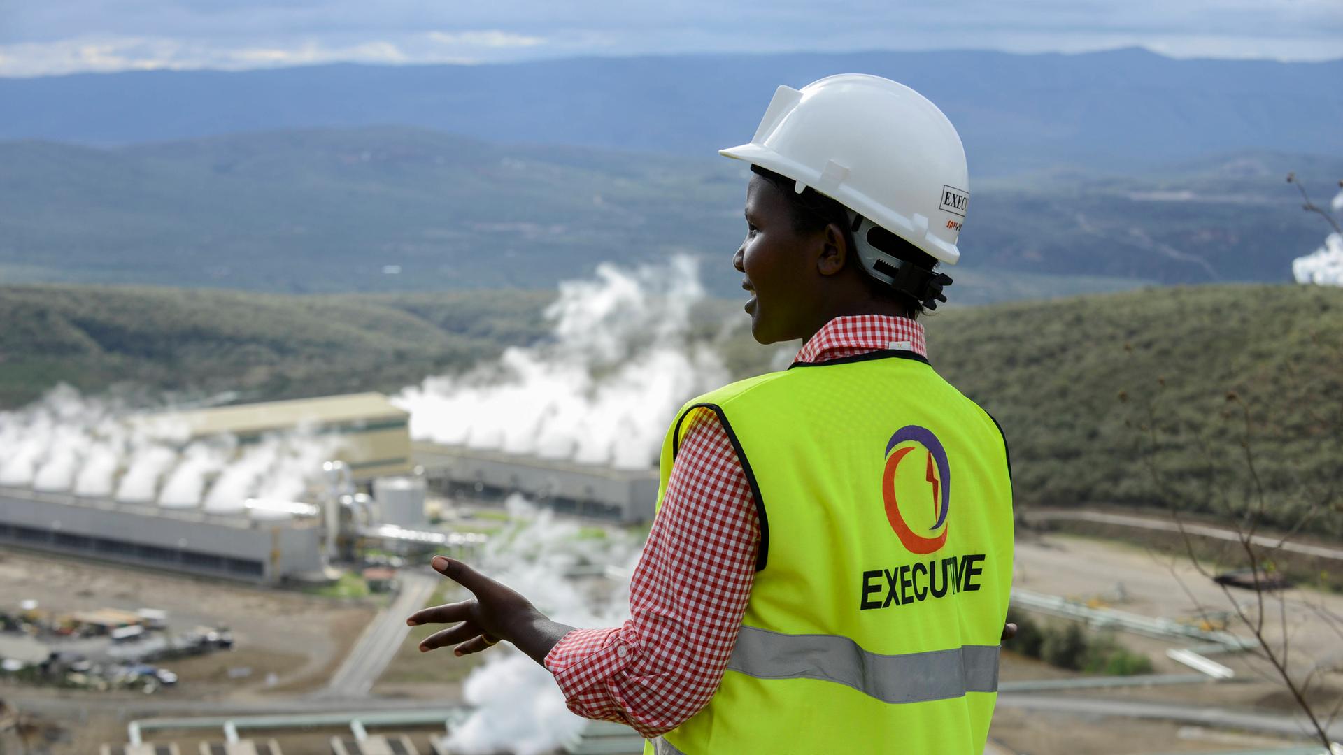 Im Vordergrund ist eine schwarze Frau mit dem Rücken zur Kamera zu sehen, im Hintergrund das Geothermie-Kraftwerk Olkaria IV in Kenia. Sie trägt einen weißen Helm, rotkariertes Hemd und darüber eine neongelbe Weste mit der Aufschrift Executive und einem Logo. 