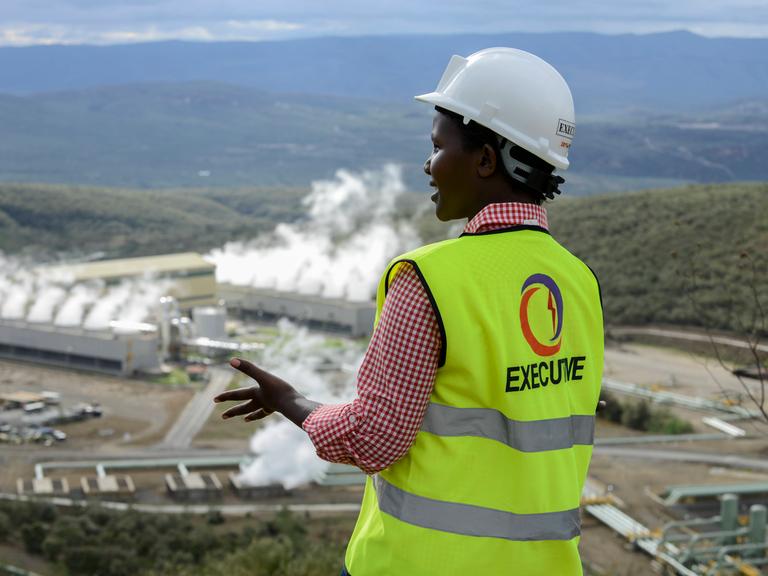 Im Vordergrund ist eine schwarze Frau mit dem Rücken zur Kamera zu sehen, im Hintergrund das Geothermie-Kraftwerk Olkaria IV in Kenia. Sie trägt einen weißen Helm, rotkariertes Hemd und darüber eine neongelbe Weste mit der Aufschrift Executive und einem Logo. 