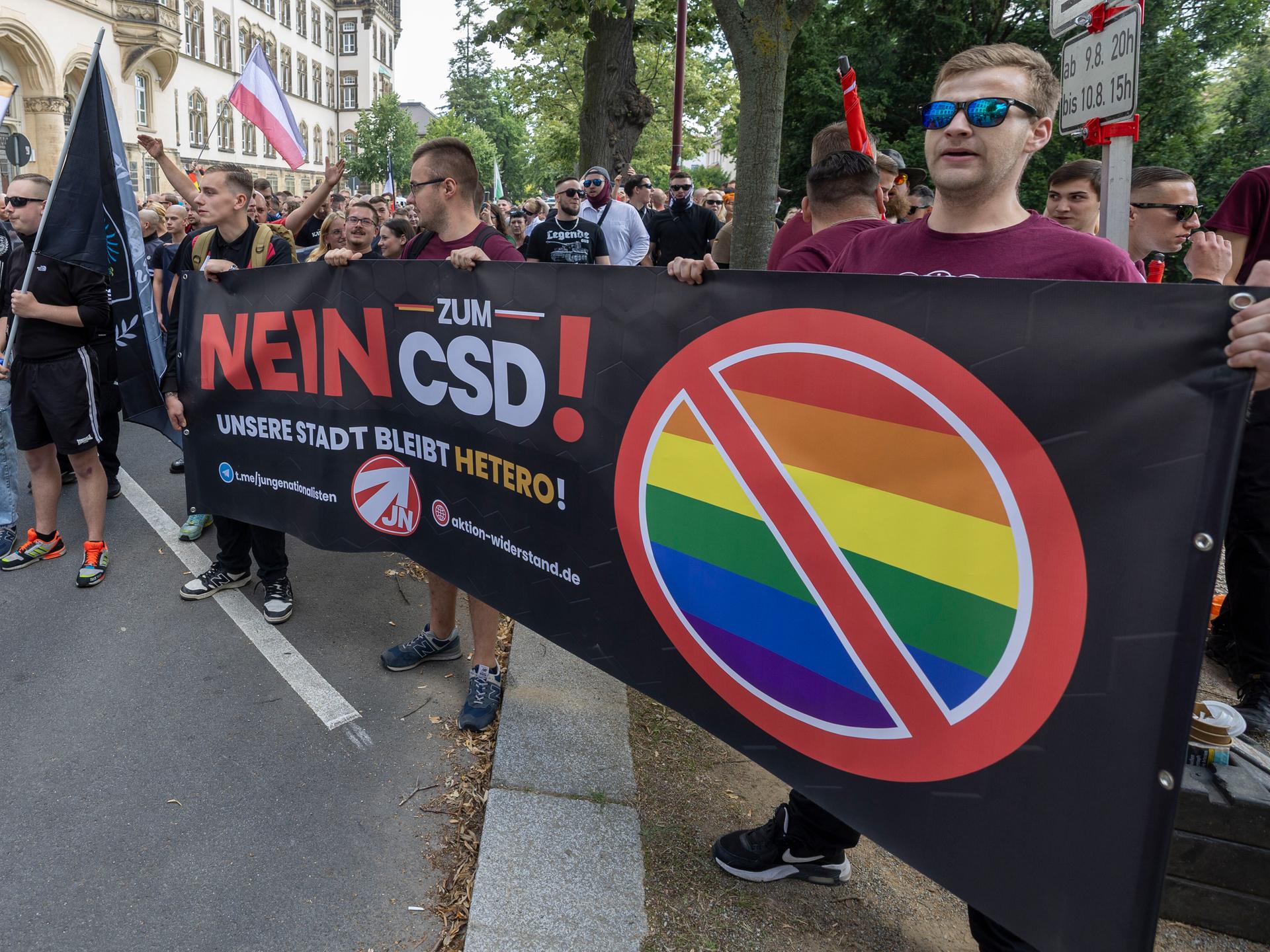 Eine Gruppe Rechtsextremer mit Sonnenbrillen hält ein Banner mit der Aufschrift "Nein zum CSD!" in die Luft. Neben dem Satz steht eine durchgestrichene Regenbogenflagge.