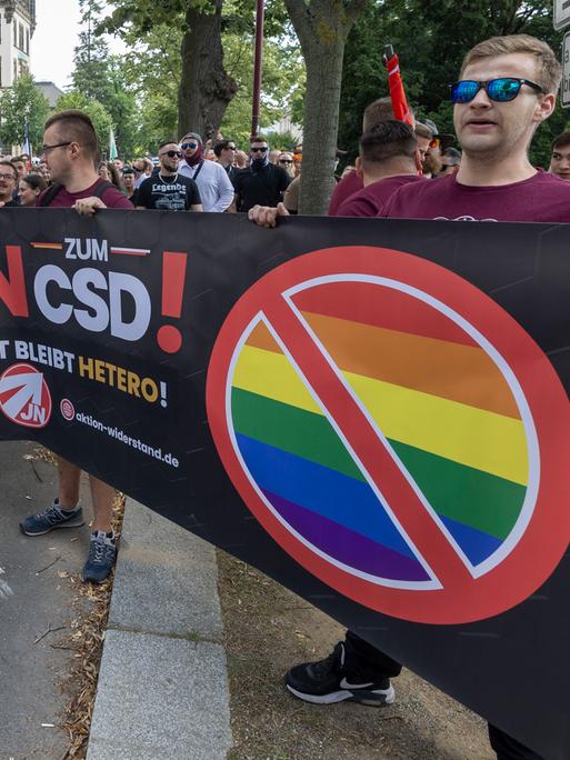 Eine Gruppe Rechtsextremer mit Sonnenbrillen hält ein Banner mit der Aufschrift "Nein zum CSD!" in die Luft. Neben dem Satz steht eine durchgestrichene Regenbogenflagge.