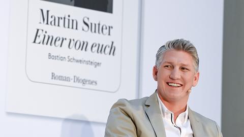 Der Fußballer Bastian Schweinsteiger sitzt in einem hellen Sakko auf einem Podium, hinter ihm an der Wand: der Buchumschlag des biografischen Romans "Einer von uns", in dem der Bestsellerautor Martin Suter aus Schweinsteigers Leben erzählt.