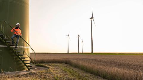 Eine Windkraftanlage. Ein Bauarbeiter steht auf einer Treppe zur Anlage, daneben Windräder auf einem Feld.