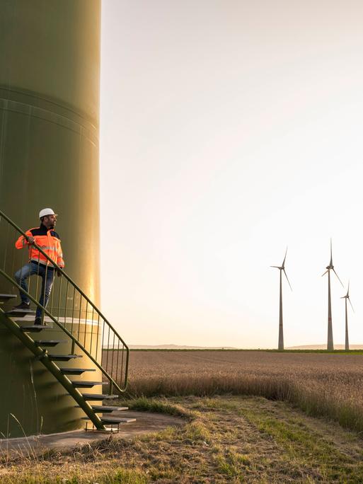 Eine Windkraftanlage. Ein Bauarbeiter steht auf einer Treppe zur Anlage, daneben Windräder auf einem Feld.