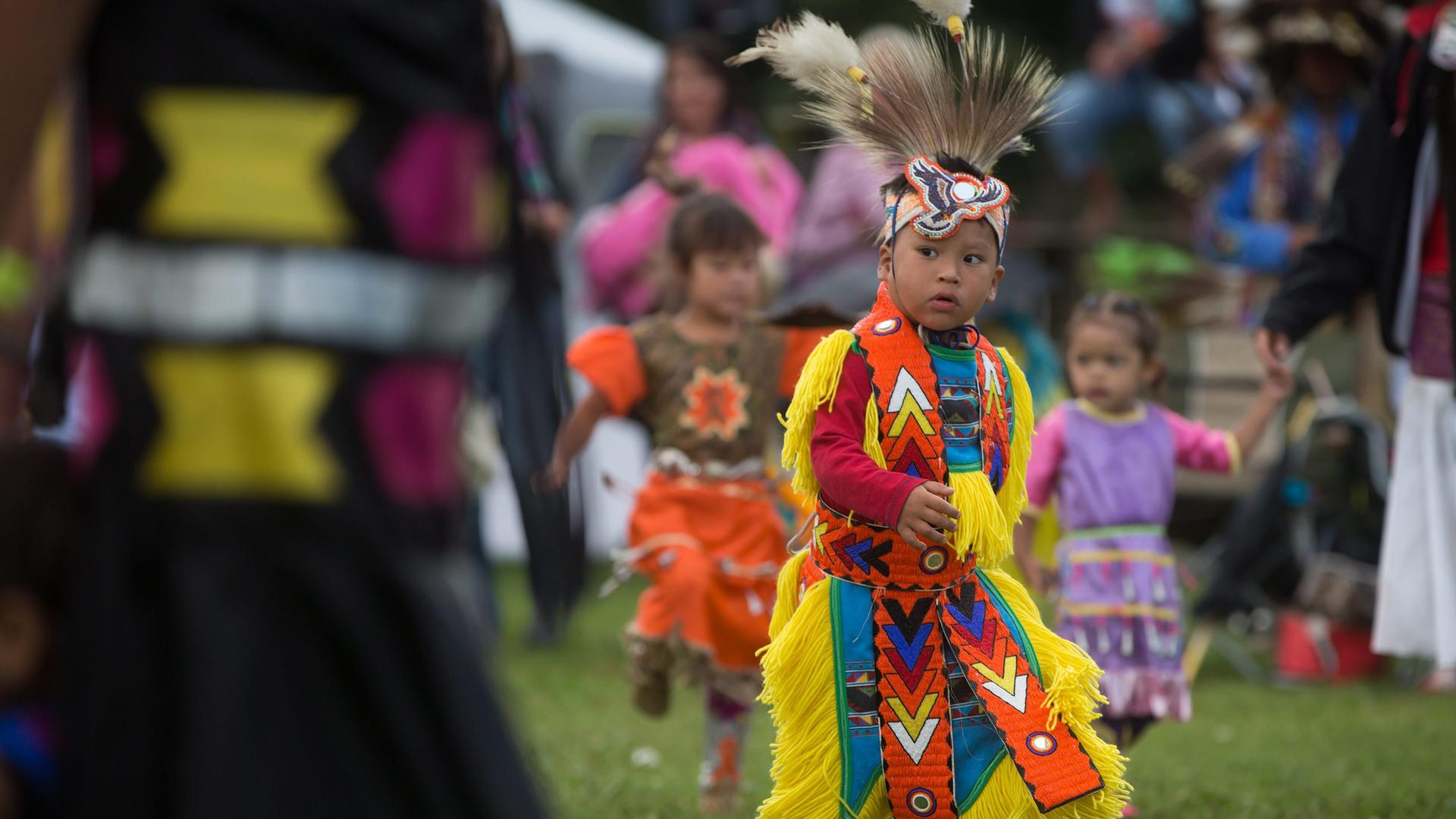 Der Junge in gelb-orangener indigener Tracht und Federn am Kopf tanzt auf einer Wiese. Dahinter unscharf zwei ebenfalls tandeznede indigene Mädchen.