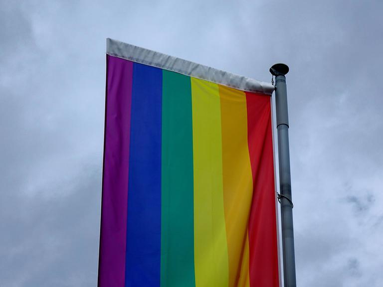 Eine Regenbogenflagge befindet sich vor dunklen Wolken. Die Fahne ist quer aufgehängt.