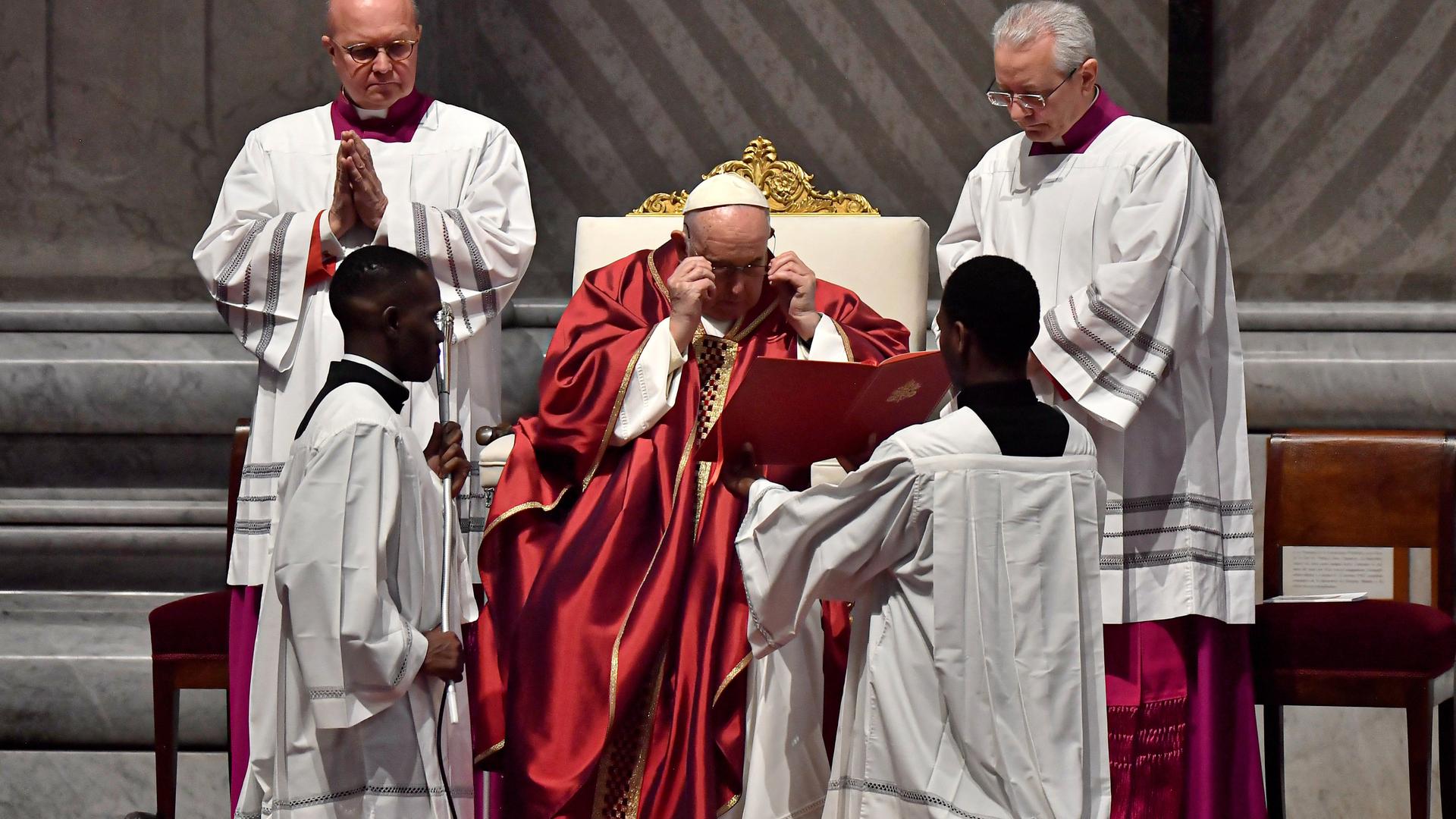 Der Papst setzt eine Brille auf, während ein Messdiener Texte vor ihm aufblättert. Der Papst setzt eine Brille auf, während ein Messdiener Texte vor ihm aufblättert.