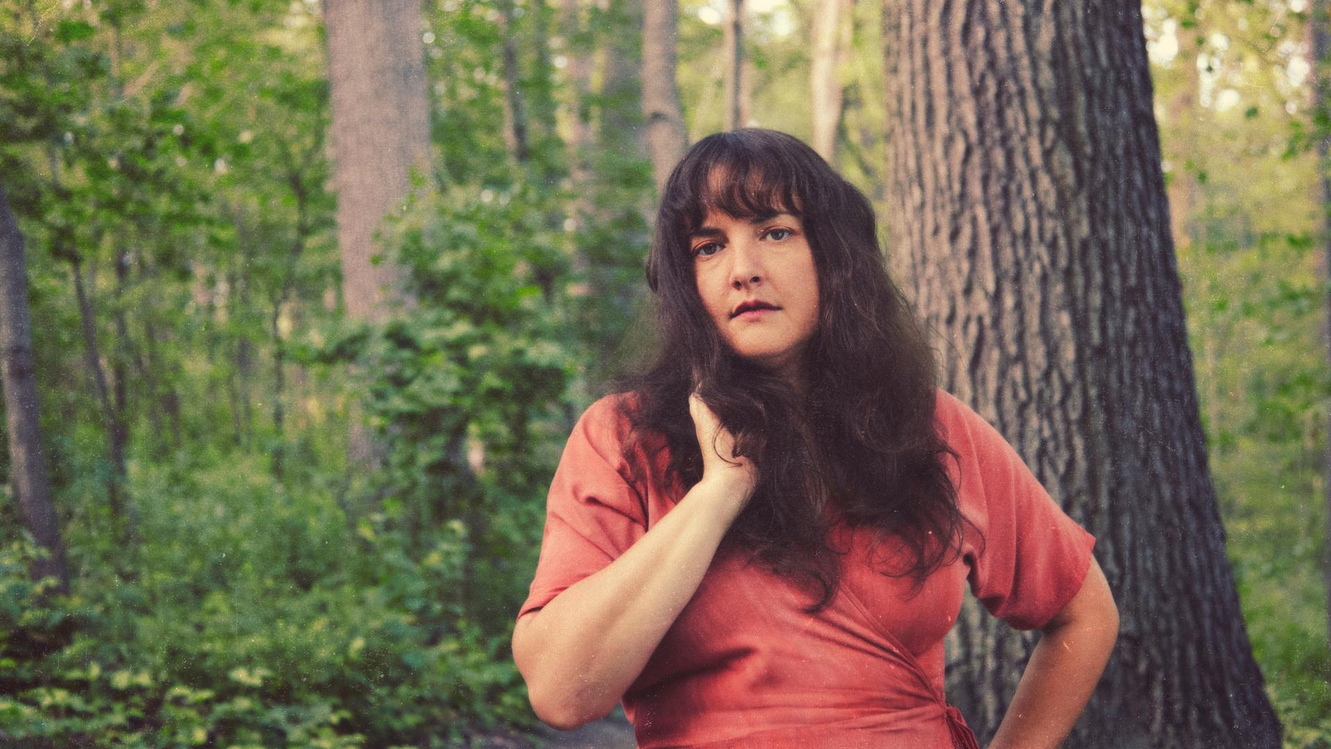 Sängerin Abigail Lapell steht in einem frischen, grünen Wald vor einem großen Baum in rotem Wickelkleid und offenen dunklen, leicht welligen Haaren.