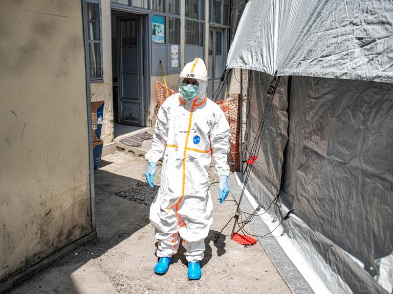 Ein Mitarbeiter reinigt ein Ebola-Behandlungszentrum in Goma (Kongo). Er trägt weiße Schutzkleidung und läuft zwischen zwei Zelten durch.