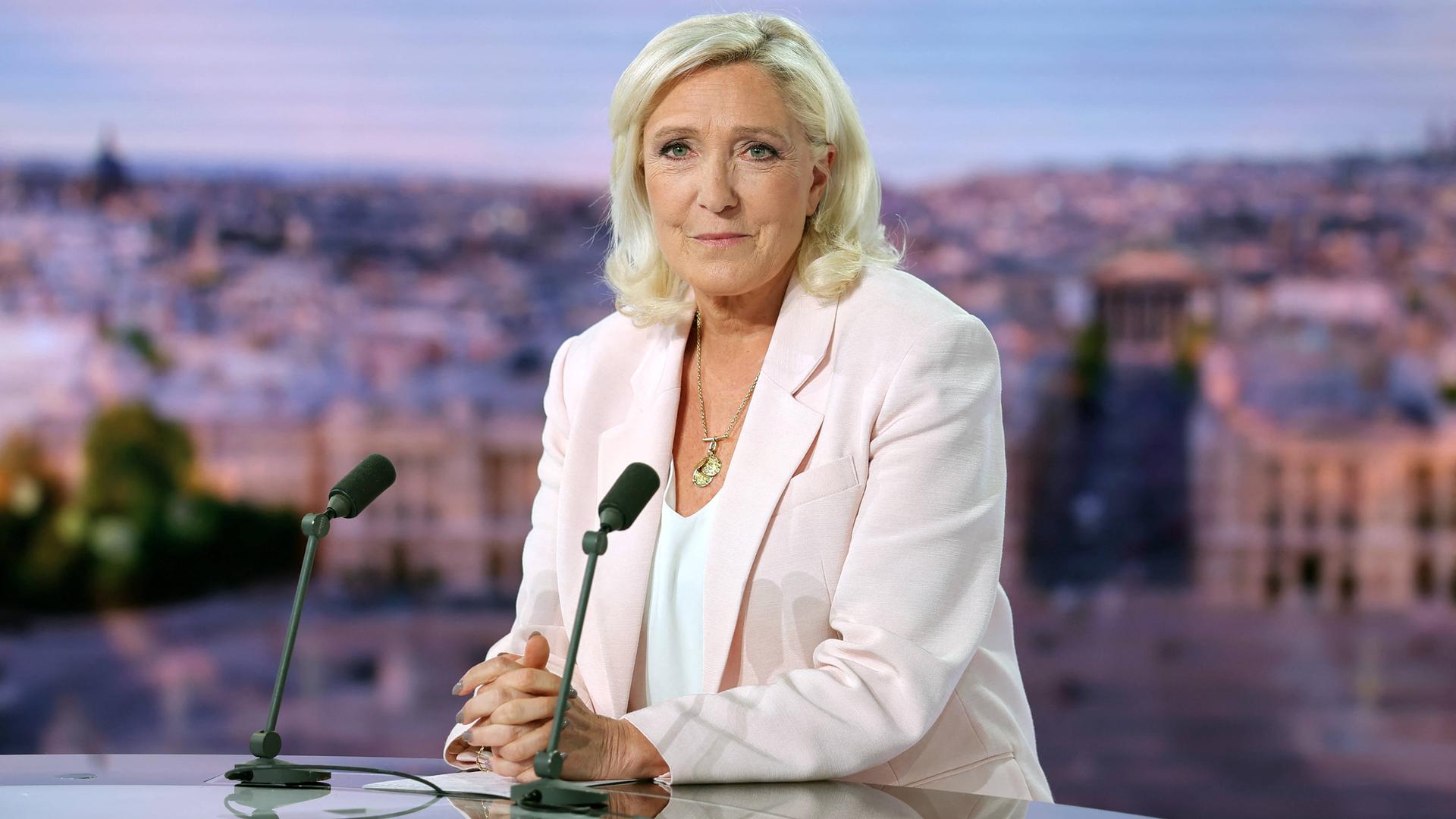 Eine blone Frau sitzt vor zwei Mikrofonen in einem Fernseh-Studio. Sie trägt einen rosanen Blazer. Eine blone Frau sitzt vor zwei Mikrofonen in einem Fernseh-Studio. Sie trägt einen rosanen Blazer.