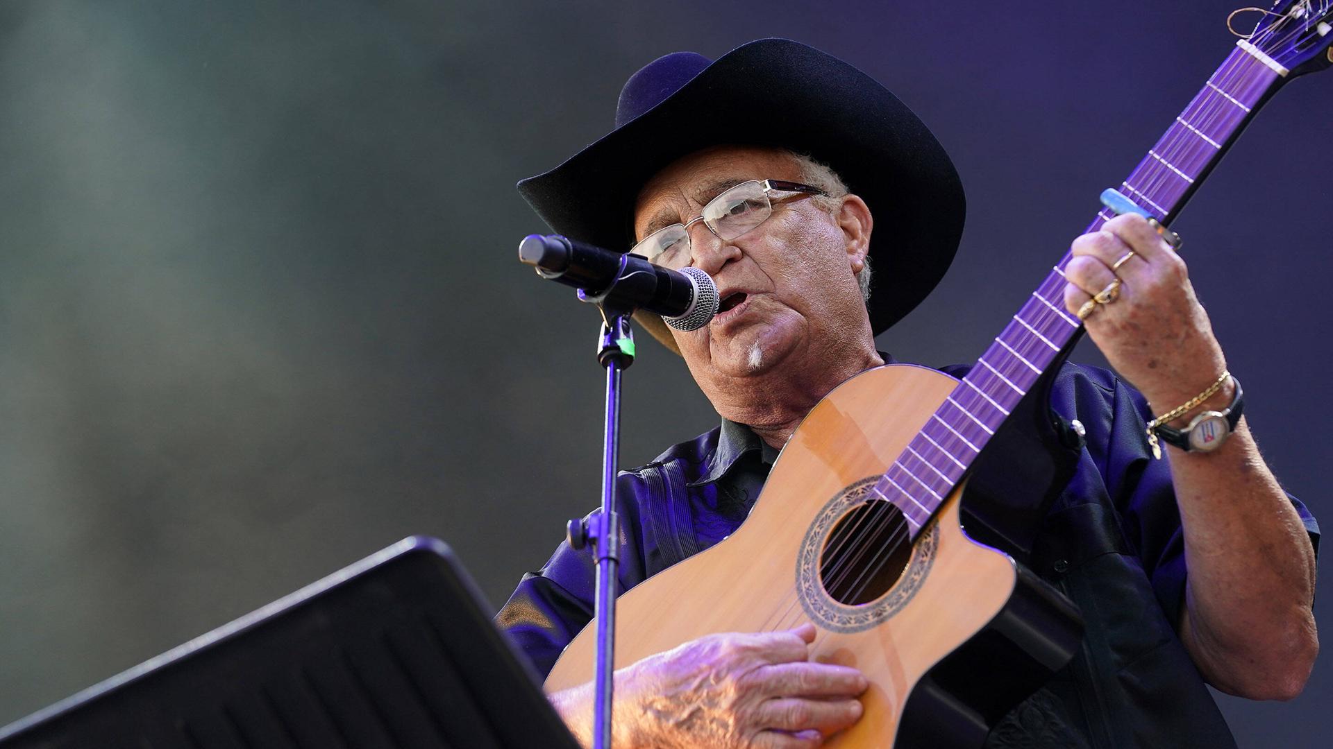 Eliades Ochoa spielt Gitarre und singt auf einer Bühne, während er einen schwarzen Hut trägt. 