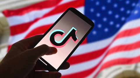 Im Vordergrund ist ein Handy mit Tik-Tok-Logo auf dem Bildschirm. Im Hintergrund sieht man die US-amerikanische Flagge.