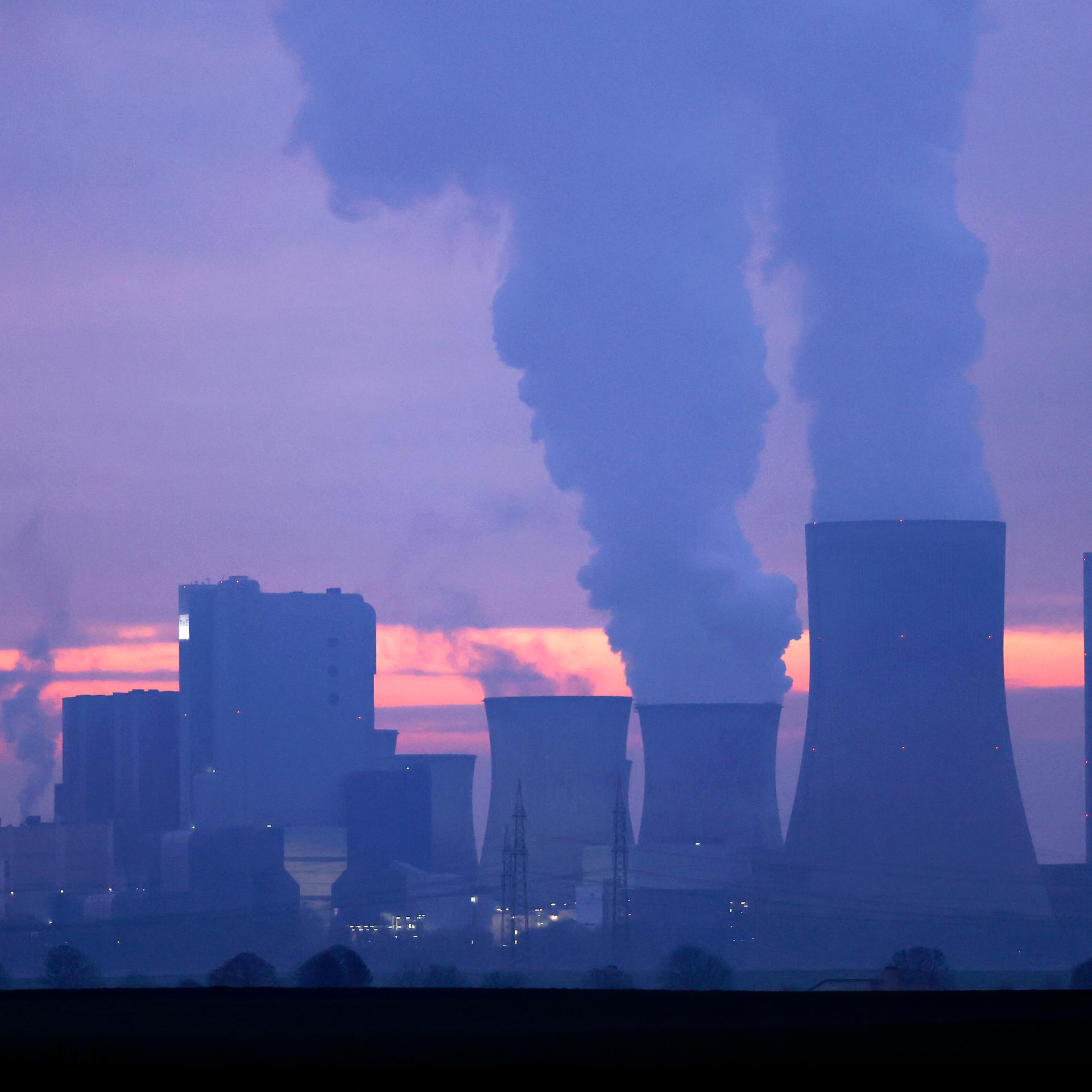 Türme eines Kohlekraftwerks stoßen Rauch aus in den abendlichen Himmel im Sonnenuntergang. 