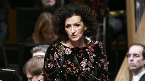 Maja Haderlap spricht auf einem Podium. Sie hat dunkelbraune Locken und trägt ein schwarzes Kleid mit Blumenstickereien.