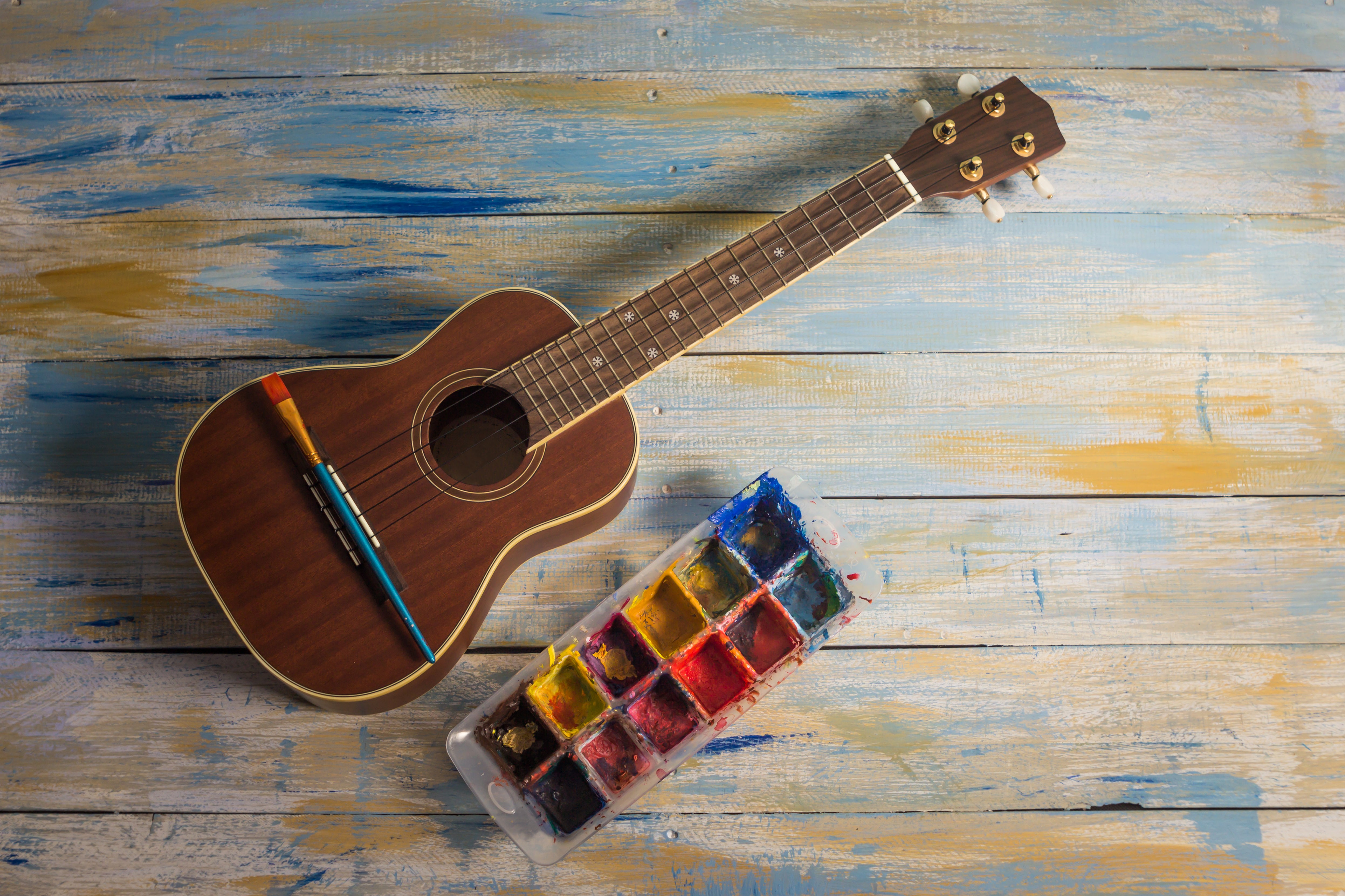 Eine Ukulele liegt neben einer Farbpalette, auf ihr sieht man einen Pinsel. Eine Ukulele liegt neben einer Farbpalette, auf ihr sieht man einen Pinsel.