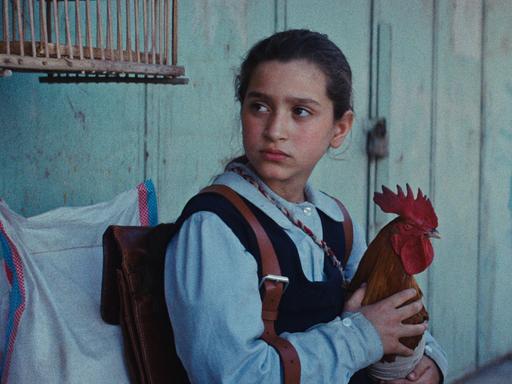 Ein Mädchen sitzt auf einer Bank und hält auf ihrem Schoß einen Hahn (Szenenbild aus dem Film "Ein Kuchen für den Präsidenten")