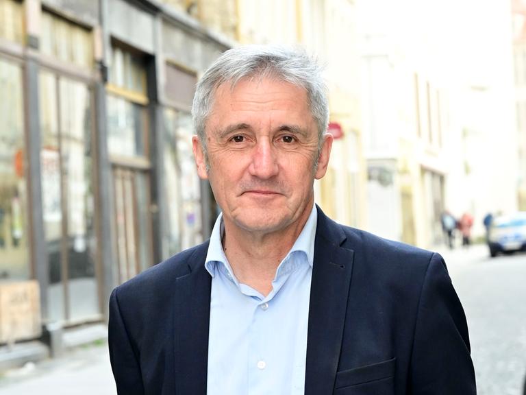 Ein Mann, Frank Richter, steht auf einer gepflasterten Straße, im Hintergrund sind Häuserfasaden zu sehen