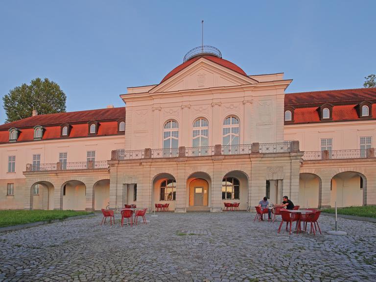 Blick auf das Deutsche Literaturarchiv in Marbach: Das Gebäude wird in rosa von der Sonne angeleuchtet, davor stehen einige Tische und Stühle. 