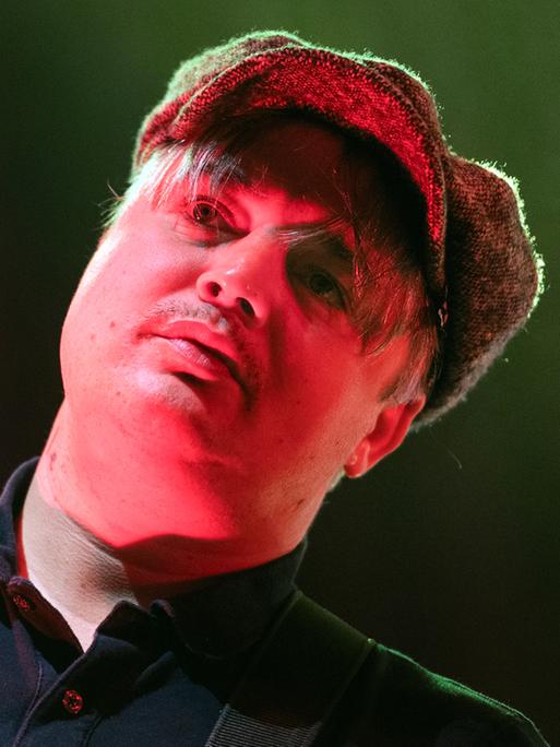 Pete Doherty mit einer Schiebermütze auf der Bühne. Sein Gesicht wird von einem Bühnenscheinwerfer rötlich angestahlt.